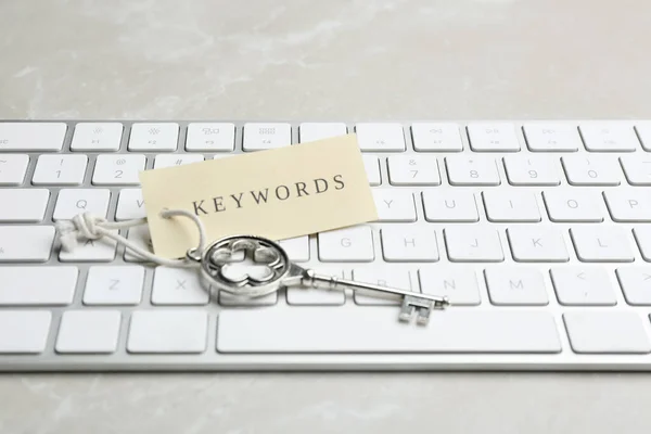 Keyboard, vintage key and tag with word KEYWORDS on light grey table, closeup