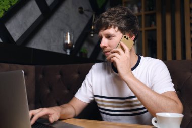 Kafedeki masada dizüstü bilgisayar kullanırken telefonda konuşan yakışıklı bir adam.