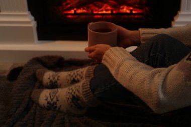 Woman in warm socks with cup of hot drink resting near fireplace at home, closeup clipart