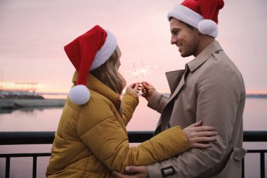 Noel Baba şapkalı bir çift nehir kenarında yanan maytaplar tutuyor.