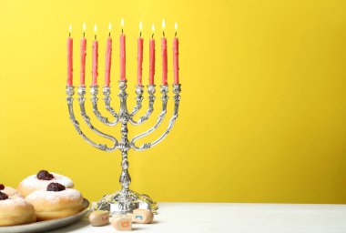 Silver menorah near sufganiyot and dreidels with symbols Nun, He, Pe, Gimel on yellow background, space for text