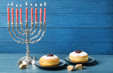 Gümüş menorah, dreidels with He, Pe, Nun, Gimel letters and sufganiyot on blue wood table, space for text. Hanuka sembolleri