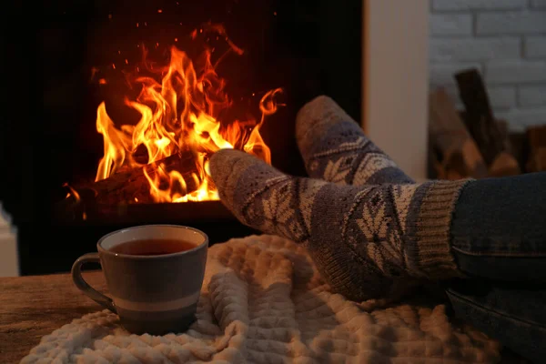 Woman in warm socks with cup of hot drink resting near fireplace at home, closeup