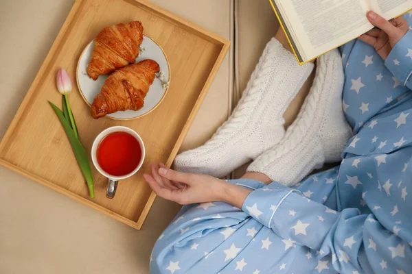 Woman with cup of tea and book resting on sofa, top view