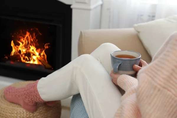 Woman with tea sitting near fireplace at home, closeup