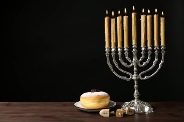 Sufganiyah yakınlarında gümüş menorah ve siyah arka planda Nun, He, Pe, Gimel sembolleri olan dreidels. Metin için boşluk