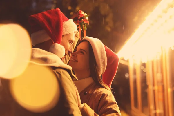 Szczęśliwa Para Kapeluszach Santa Stojących Pod Jemiołą Kilka Zewnątrz Efekt — Zdjęcie stockowe