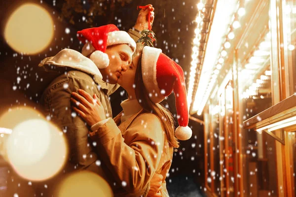 Happy couple kissing under mistletoe bunch outdoors in snowy evening, bokeh effect