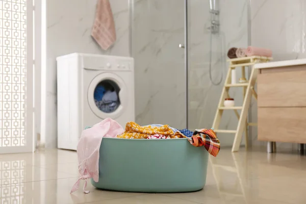 Plastic laundry basket full of different clothes on floor in bathroom