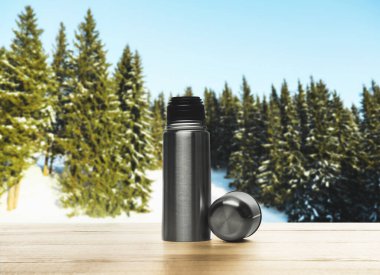 Karlı ormandaki ahşap masada termos şişesi.