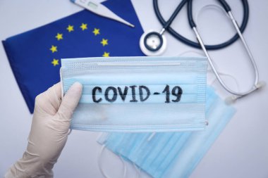 Tıbbi eşyalar ve Avrupa Birliği bayrağı üzerinde COVID-19 sözcüğüyle maskeli doktor, yakın plan
