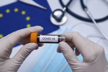 Tıbbi eşyalar ve Avrupa Birliği bayrağı üzerinde COVID-19 etiketli örnek tüp tutan doktor, yakın plan