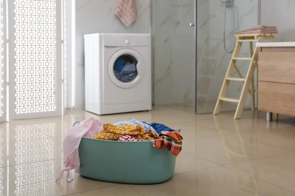 Plastic laundry basket full of different clothes on floor in bathroom