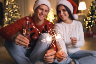 Noel Baba şapkalı çift ellerinde ışıltı ve şampanya bardakları, havai fişeklere odaklan. Noel kutlaması