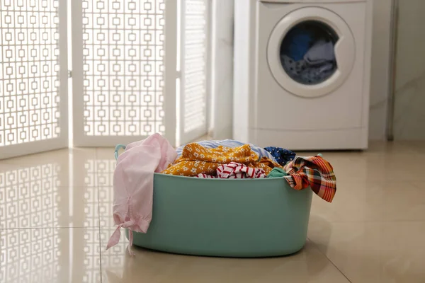 Plastic laundry basket full of different clothes on floor in bathroom