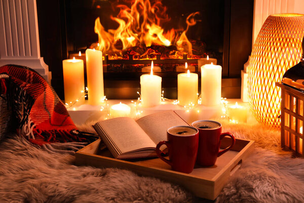 Cups of hot drink and open book near fireplace at home