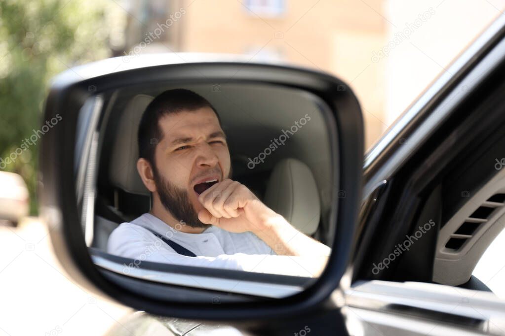 Tired man yawning in his modern car, view through car side mirror