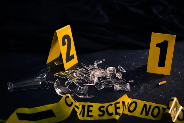 Broken bottle, yellow tape and evidence markers on black slate table. Crime scene