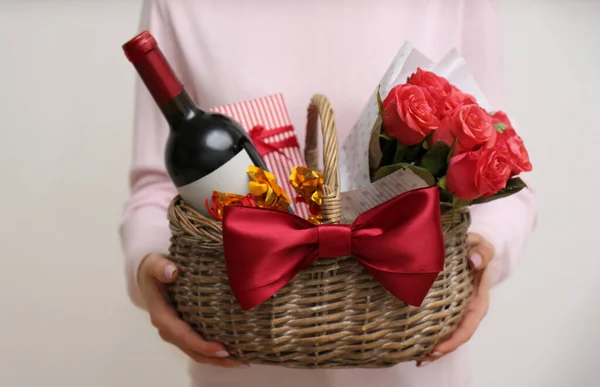 Woman holding wicker basket with gift, bouquet and wine on light grey background, closeup