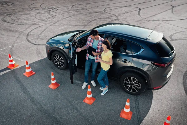 Instructor with clipboard near car and his student outdoors, above view. Driving school exam