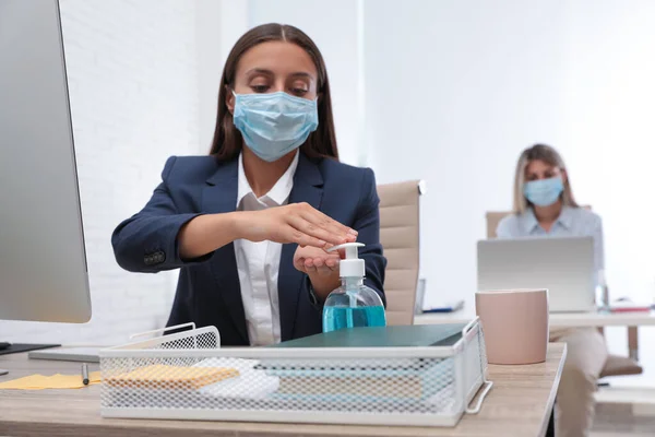 Office employee in mask applying hand sanitizer at workplace