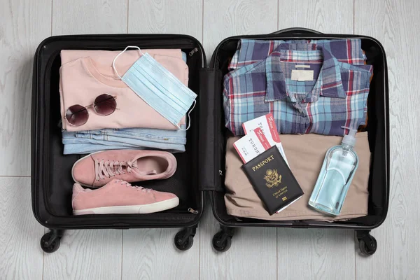 Open suitcase with passport, tickets, sanitizer and protective mask on wooden floor, top view. Travel during quarantine