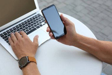 Akıllı telefonlu bir adam açık kafede dizüstü bilgisayarla çalışıyor.