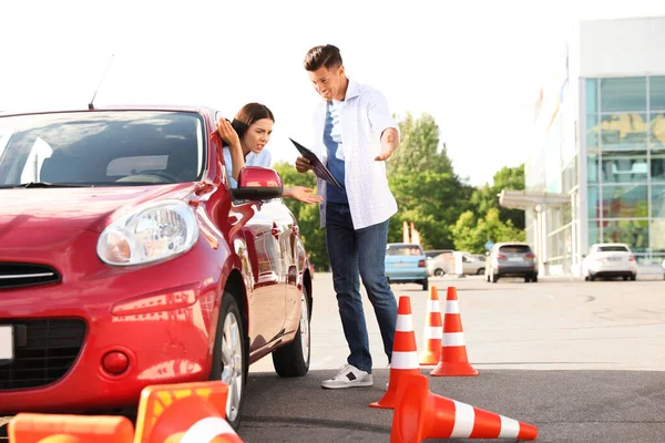 Stressed young woman in car near instructor and fallen traffic cones outdoors. Failed driving school exam