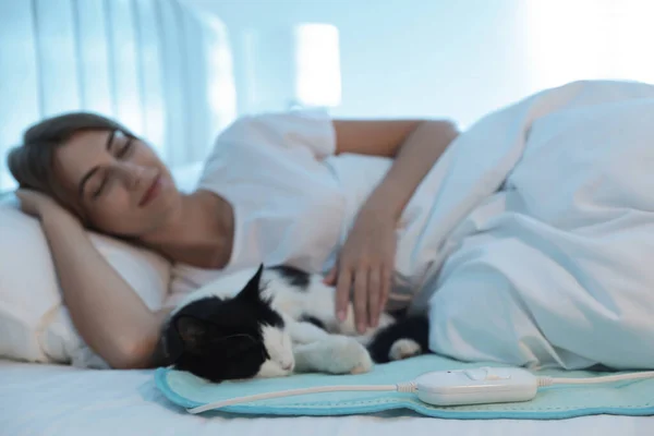 Young woman and cat in bed with electric heating pad, focus on cable