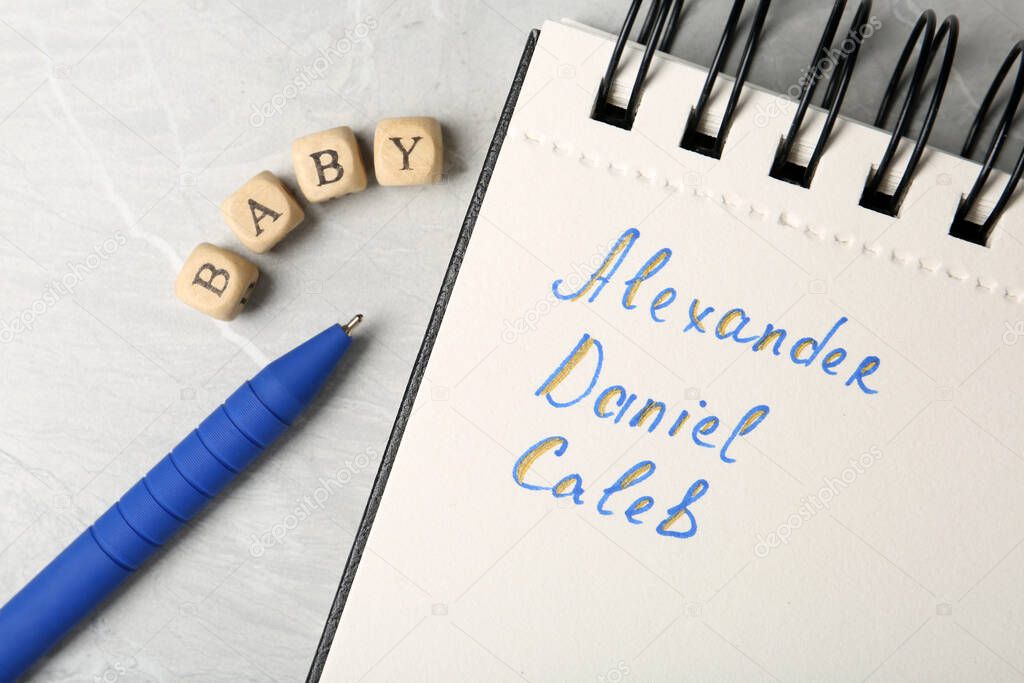 Notebook with different baby names, pen and wooden cubes on grey marble table, flat lay