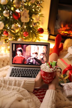 MYKOLAIV, UKRAINE - 23 Aralık 2020: Harry Potter ve Filozof Stone 'un filmini evinde şöminenin yanındaki dizüstü bilgisayarda izleyen tatlı içkili kadın, yakın plan. Sıcak kış tatili atmosferi