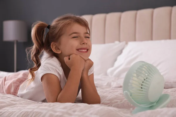 小さな女の子が部屋のベッドでポータブルファンからの空気の流れを楽しんでいます 夏の暑さ — ストック写真