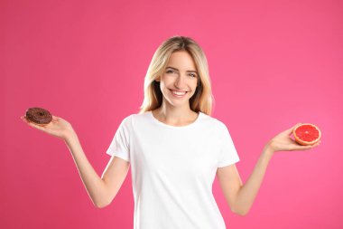 Pembe arka planda donut ve sağlıklı greyfurt arasında seçim yapan bir kadın.