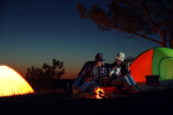 Couple with flashlight reading book near bonfire at night. Camping season