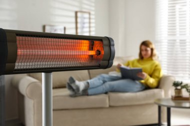 Oturma odasında kitap okuyan kadın, elektrikli kızılötesi ısıtıcıya odaklan.