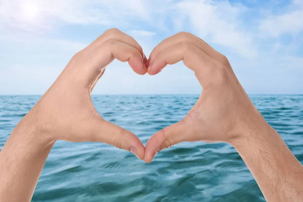 Man making heart with hands near sea on sunny day, closeup