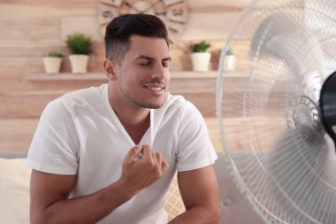 Adam oturma odasındaki kanepede vantilatörden hava akışının keyfini çıkarıyor. Yaz sıcağı