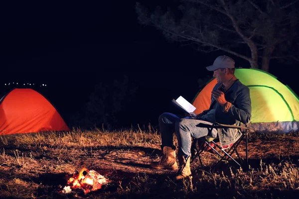 Man with flashlight reading book near bonfire at night. Camping season