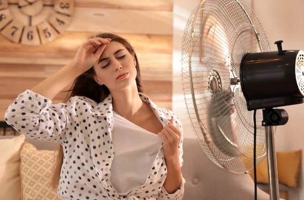 Woman enjoying air flow from fan on sofa in living room. Summer heat