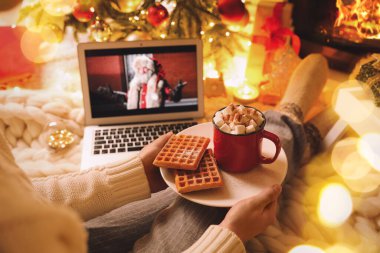 MYKOLAIV, UKRAINE - 23 Aralık 2020: Kakaolu ve gofretli kadın şöminenin yanındaki dizüstü bilgisayarda The Christmas Chronicles filmini izliyor, yakın plan. Sıcak kış tatili atmosferi
