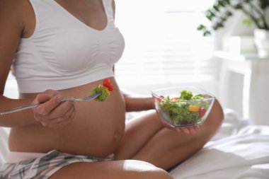 Yatak odasında bir kase sebze salatasıyla genç hamile kadın, yakın plan. Bebek sağlığıyla ilgilenmek.
