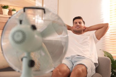 Adam oturma odasındaki kanepede vantilatörden hava akışının keyfini çıkarıyor. Yaz sıcağı