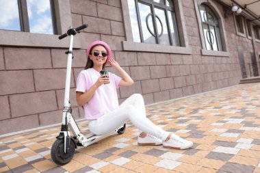 Bir fincan kahveli genç kadın, dışarıda elektrikli scooter 'ın üzerinde oturuyor.