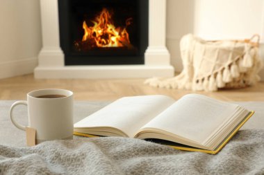 Cup of hot tea and book near fireplace at home. Cozy atmosphere clipart