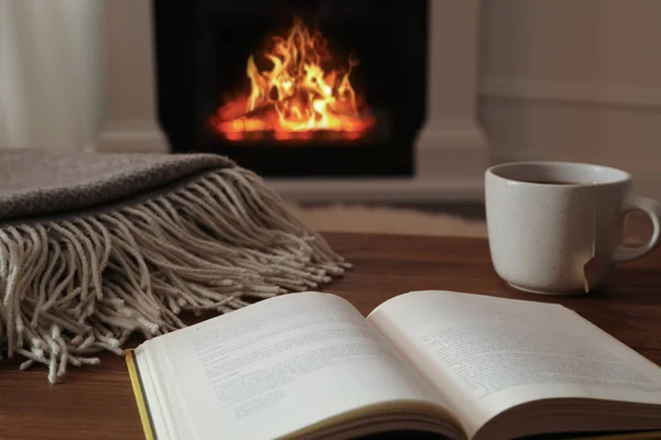 Cup of hot tea and book on wooden table near fireplace at home. Cozy atmosphere