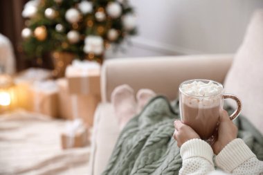 Woman with delicious hot drink wrapped in plaid on sofa at home, closeup. Space for text clipart