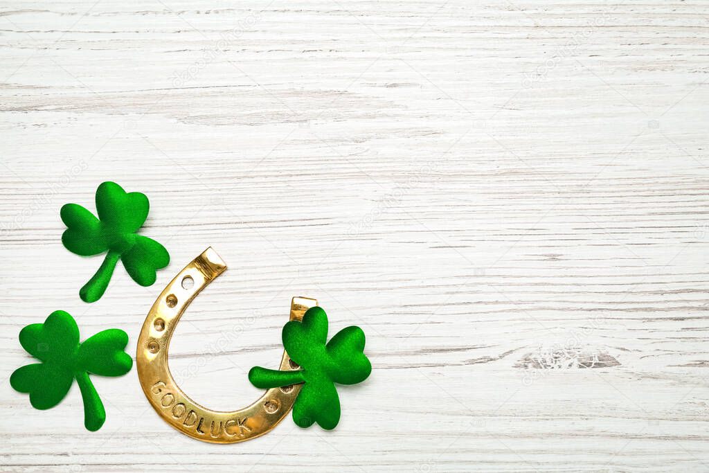 Golden horseshoe and decorative clover leaves on white wooden table, flat lay with space for text. Saint Patrick's Day celebration