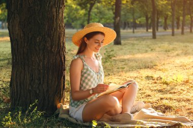 Parktaki ağacın yakınındaki yeşil çimenler üzerine kitap okuyan genç bir kadın.