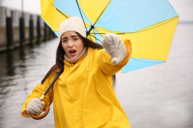 Sarı yağmurluk giymiş, şemsiyeli kadın nehrin yanında rüzgarın esintisine kapılmış.