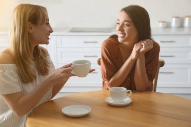 Young women talking while drinking tea at table in kitchen clipart